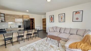A modern living room with a sofa, coffee table, and chairs. There is a kitchen area with cabinets and appliances.