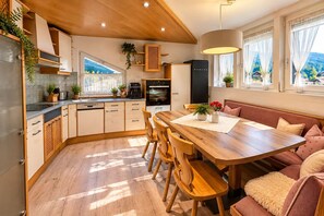 Eine moderne Küche mit Holzschränken, eine Essecke mit Holztisch und -stühlen sowie ein Blick ins Freie durch große Fenster.
