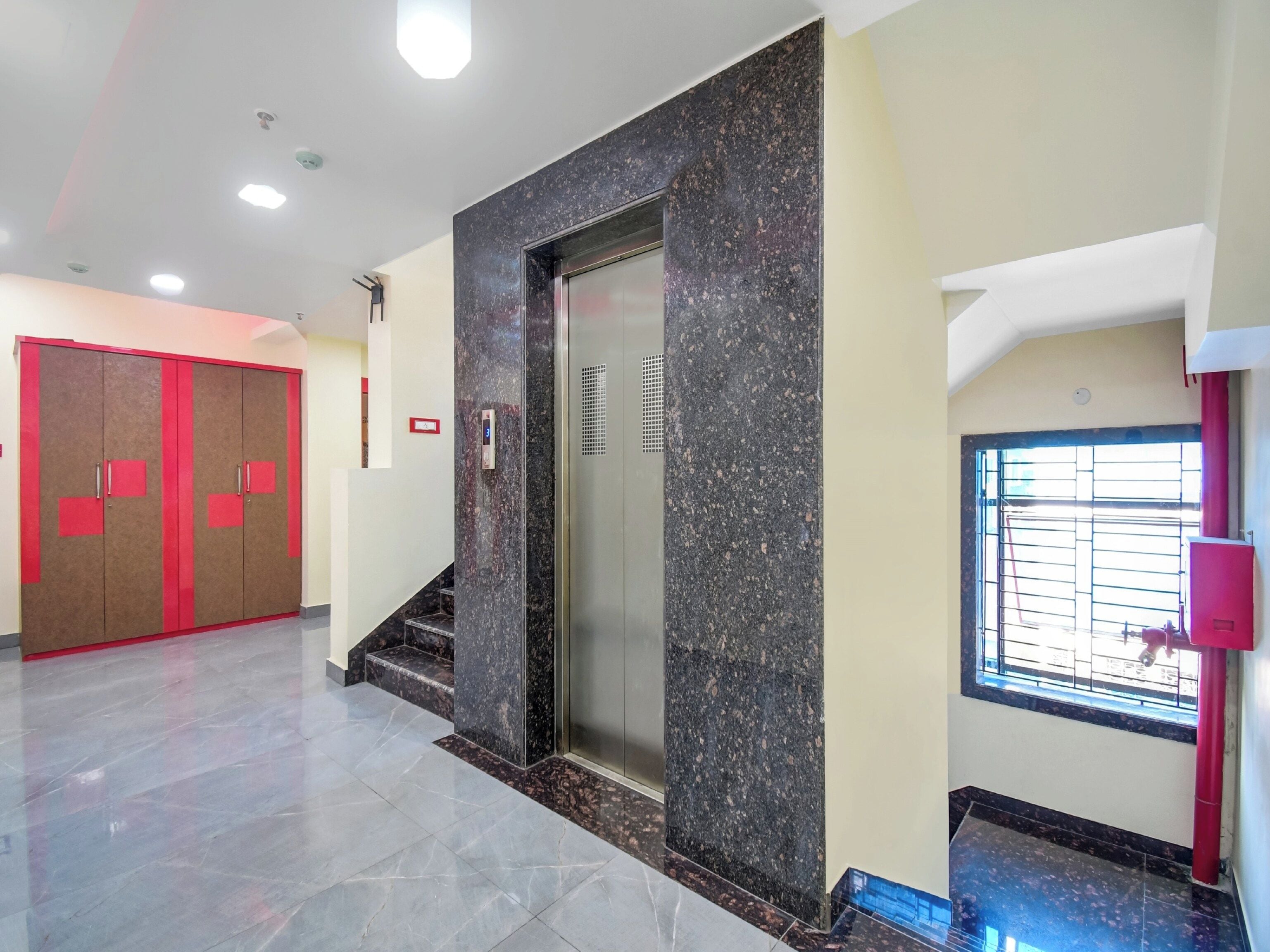 A hotel lobby with a marble floor, a red and brown wardrobe, a marble elevator, and a window with a metal grille.