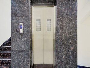 An elevator with a digital display and a control panel, surrounded by granite walls and a staircase. - Hotel O THE CROWN COMFORTS (Bidhannagar)