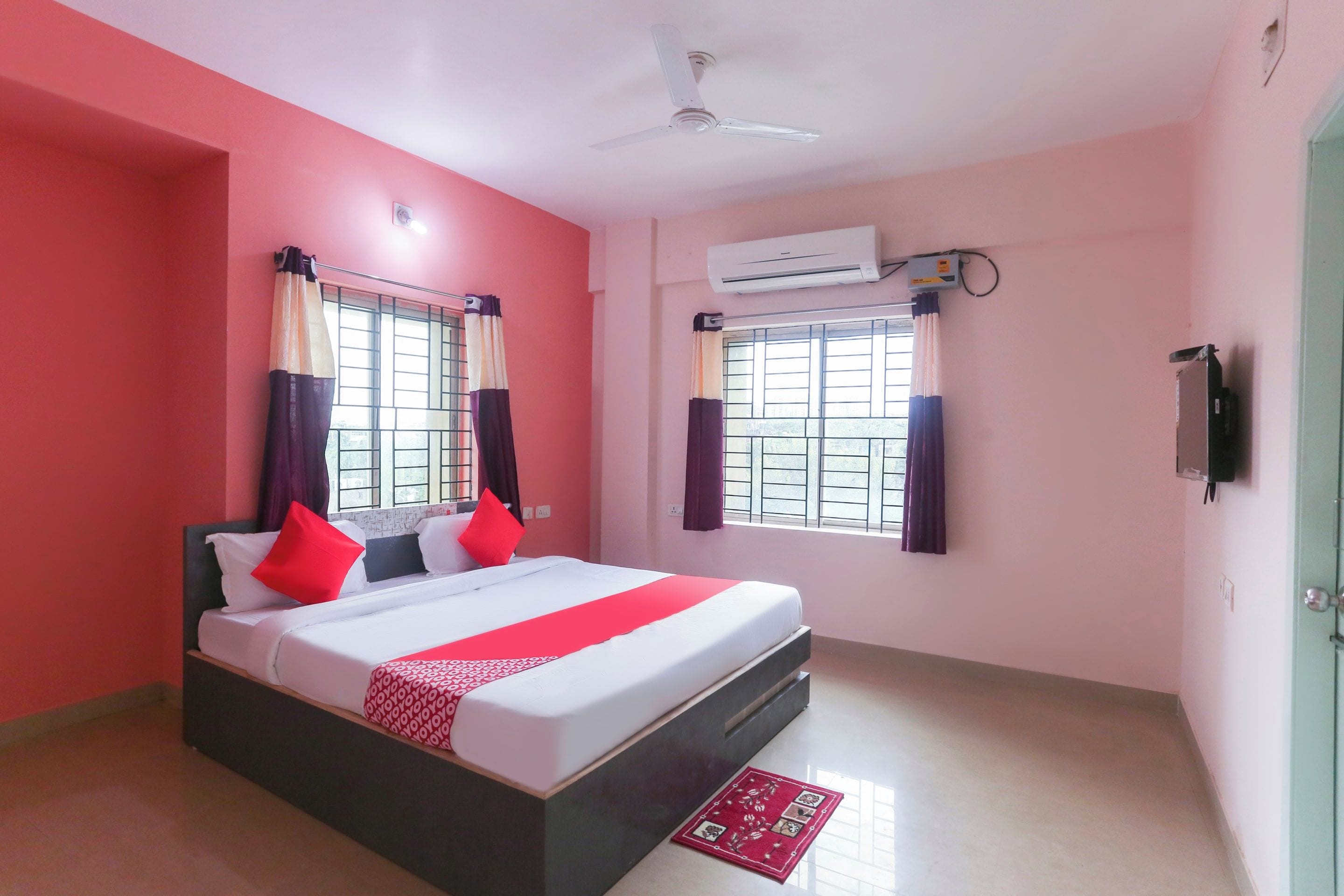 A hotel room with a bed, a ceiling fan, an air conditioning unit, and a window with curtains.