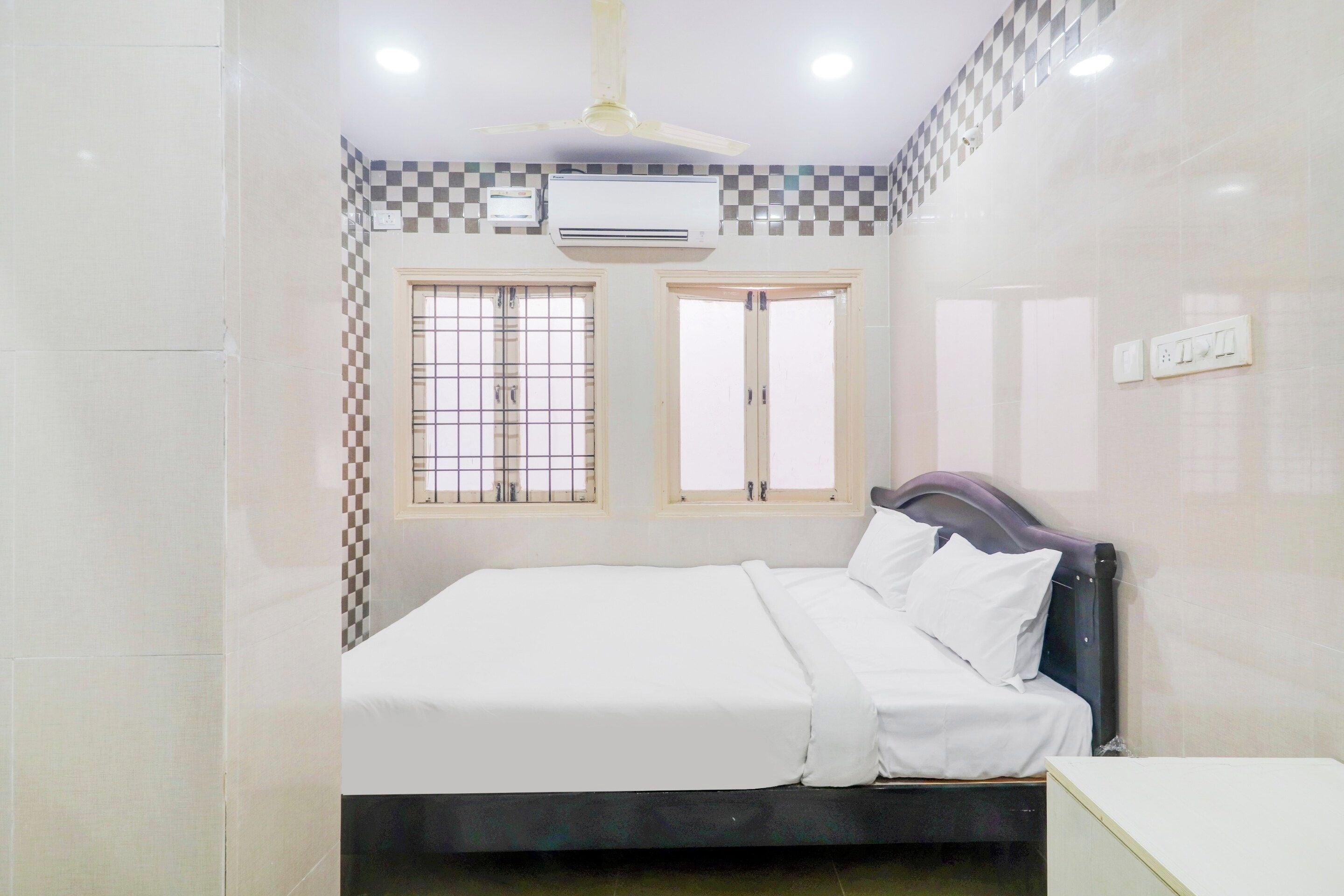 A bedroom with a bed, an air conditioning unit, two windows with bars, and a ceiling fan.