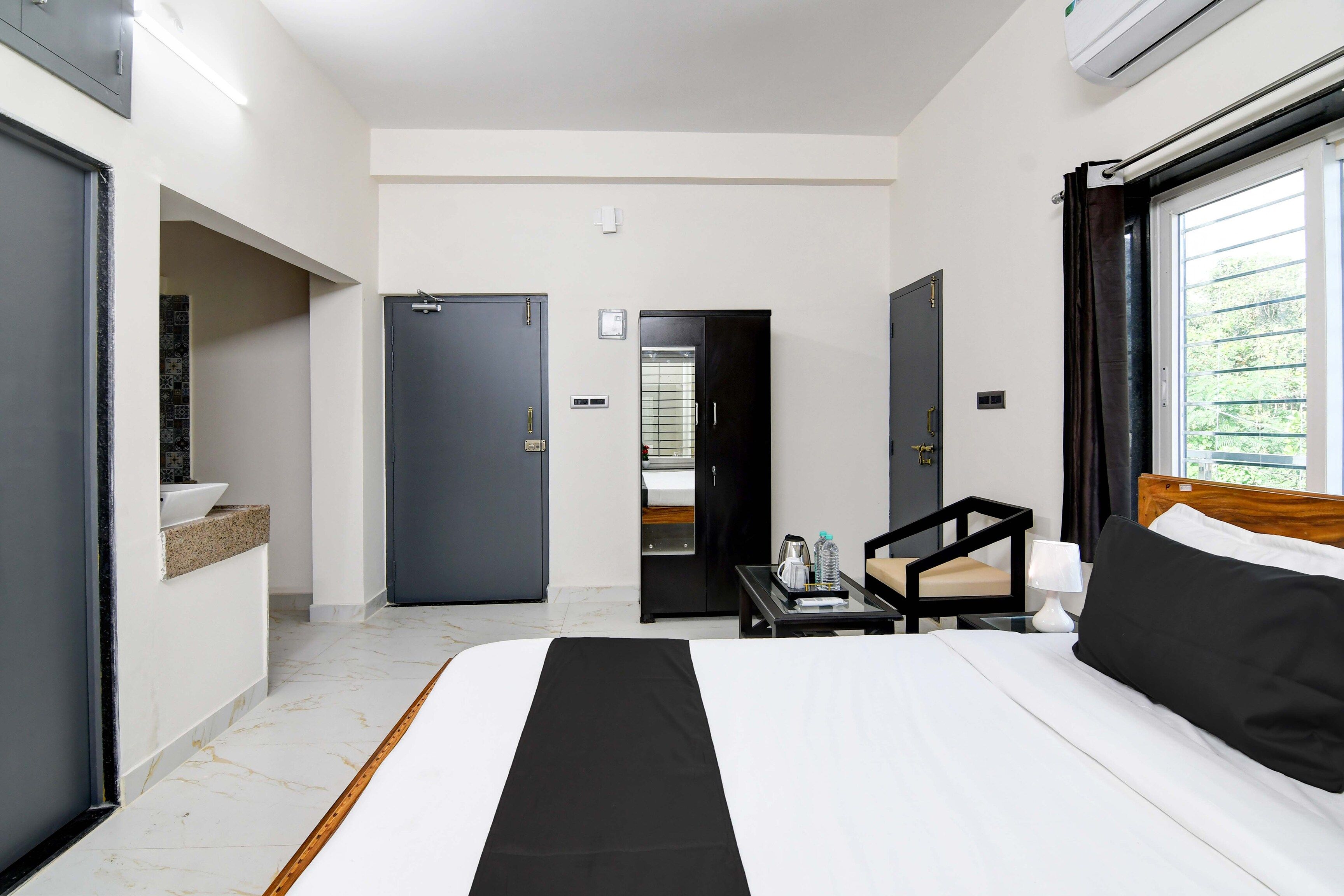 A hotel room with a bed, a small table with a lamp, a chair, and a view of greenery outside.