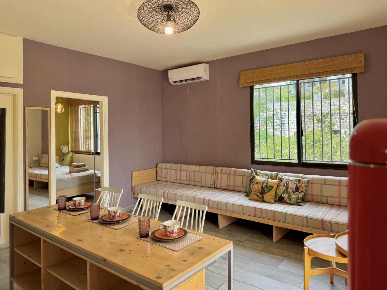 Una habitación con una mesa de comedor de madera, un sofá con cojines, un espejo y una ventana con vista a la vegetación.