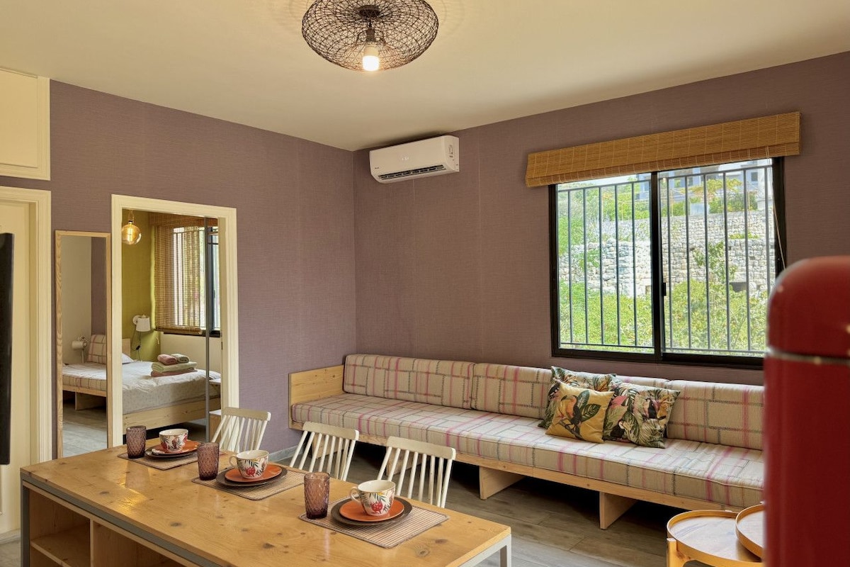 Quarto com mesa de jantar de madeira, sofá com almofadas, espelho e janela com vista para o verde.