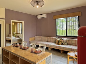 A room with a wooden dining table, a sofa with cushions, a mirror, and a window with a view of greenery. - Beit El Berbara by Stayinn (Berbara)