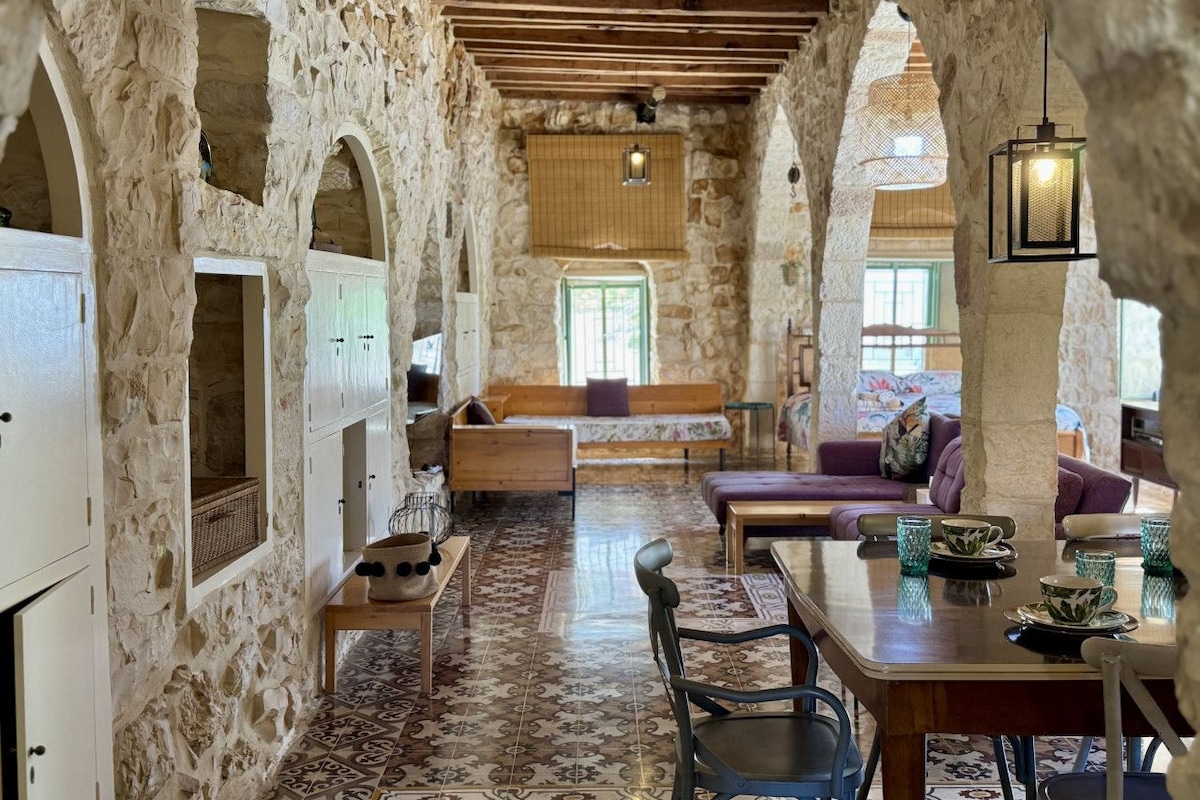 Interior rústico com paredes de pedra, vigas de madeira, área de estar central e mesa de jantar com cadeiras.