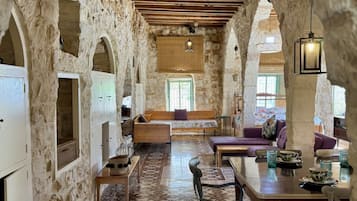 A rustic interior with stone walls, wooden beams, a central seating area, and a dining table with chairs.