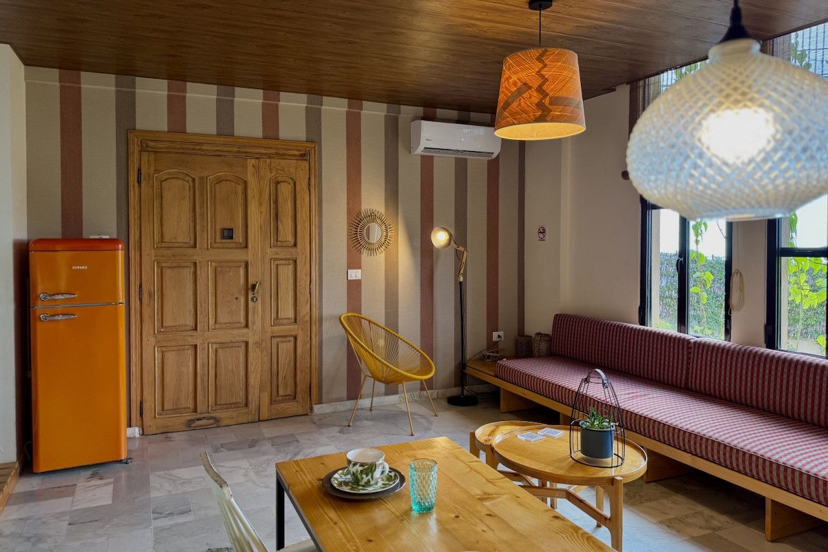 Quarto acolhedor com mesa de jantar de madeira, um sofá e uma cadeira. Há uma porta de madeira e um aparelho de ar condicionado na parede.