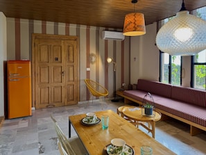 Quarto acolhedor com mesa de jantar de madeira, um sofá e uma cadeira. Há uma porta de madeira e um aparelho de ar condicionado na parede.