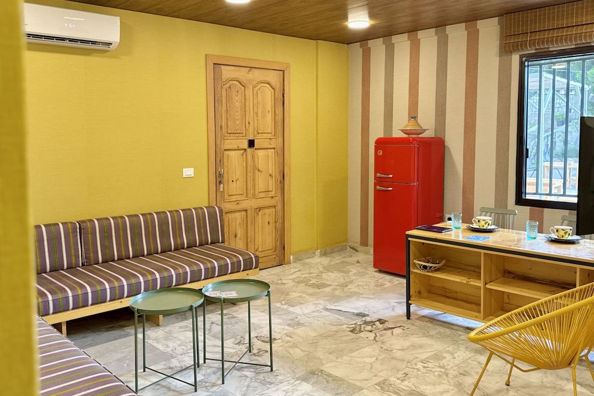 Quarto compacto com sofá, mesa pequena, geladeira vermelha e janela com vista.