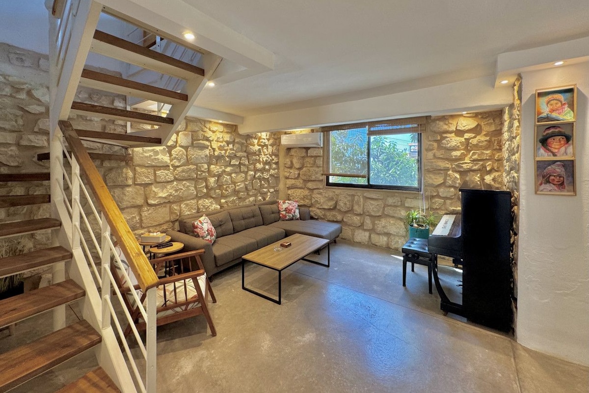 Uma sala de estar ampla com parede de pedra, escada de madeira, sofá, mesa de centro e piano.