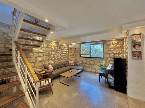 A spacious living area with a stone wall, a wooden staircase, a sofa, a coffee table, and a piano. - Beit El Berbara by Stayinn (Berbara)