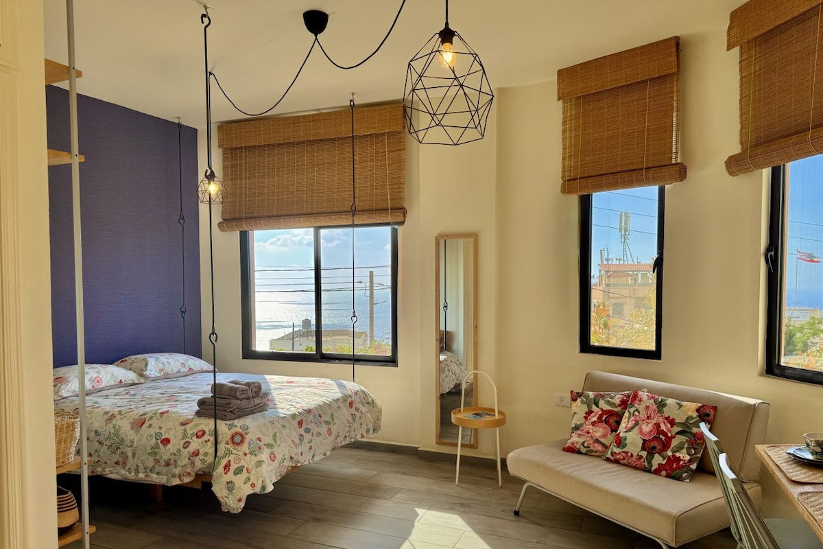 Quarto com cama, sofá, poltrona, espelho e janela com vista.
