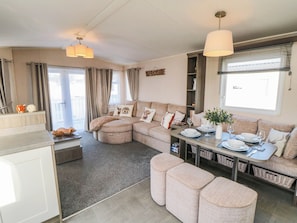 A modern living room with a sectional sofa, ottomans, a dining table set for four, and a large window with blinds. - Gosling 14 (Scarborough)