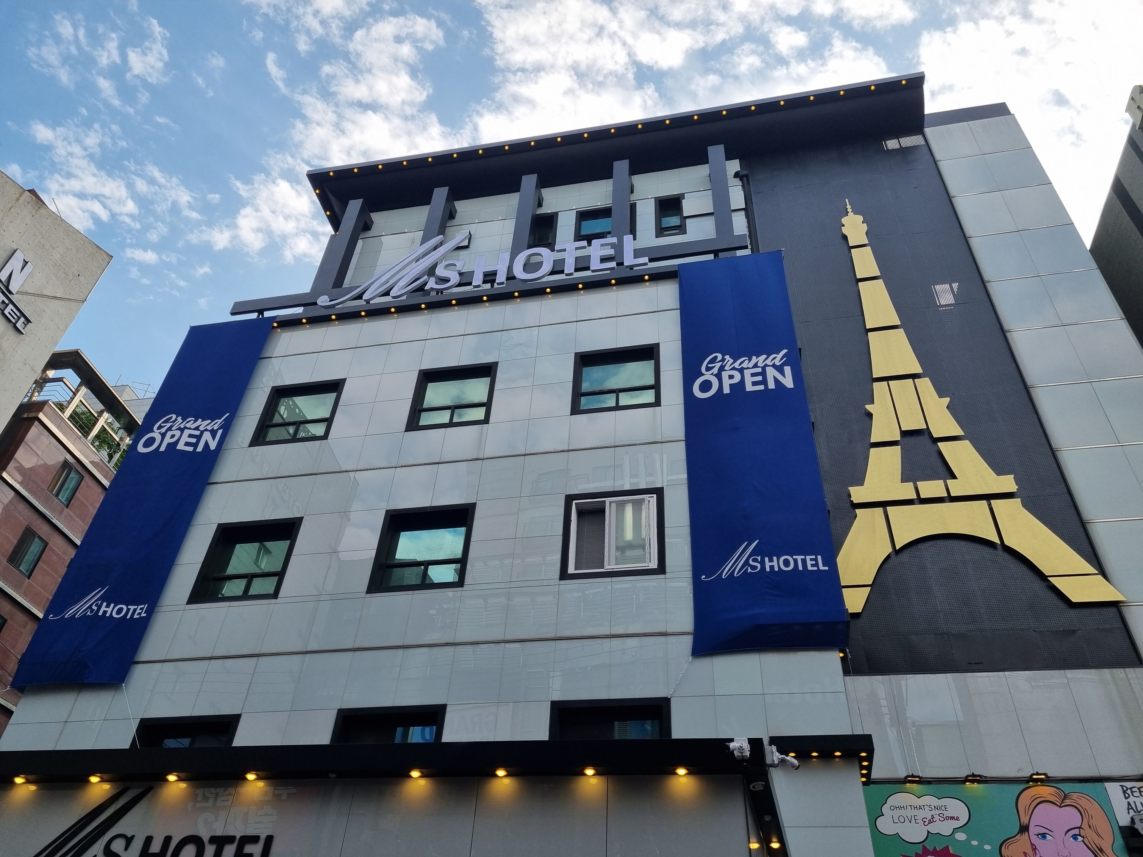Un edificio de hotel con un cartel llamativo que dice 'MS HOTEL' y un gran banderín azul que anuncia 'Gran APERTURA'.