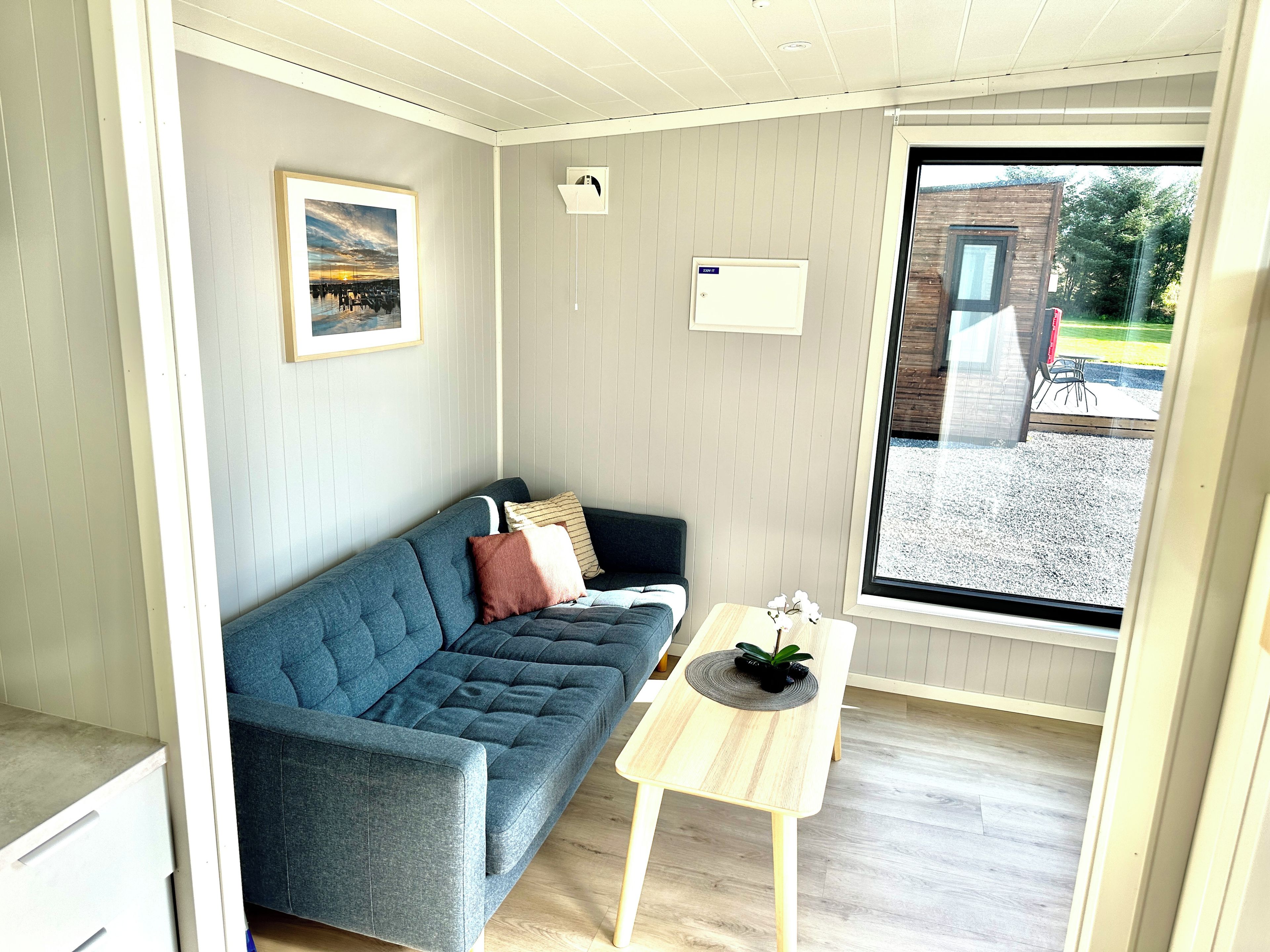 A room with a blue sofa, a small wooden table, a framed picture on the wall, and a window with a view of the outside.