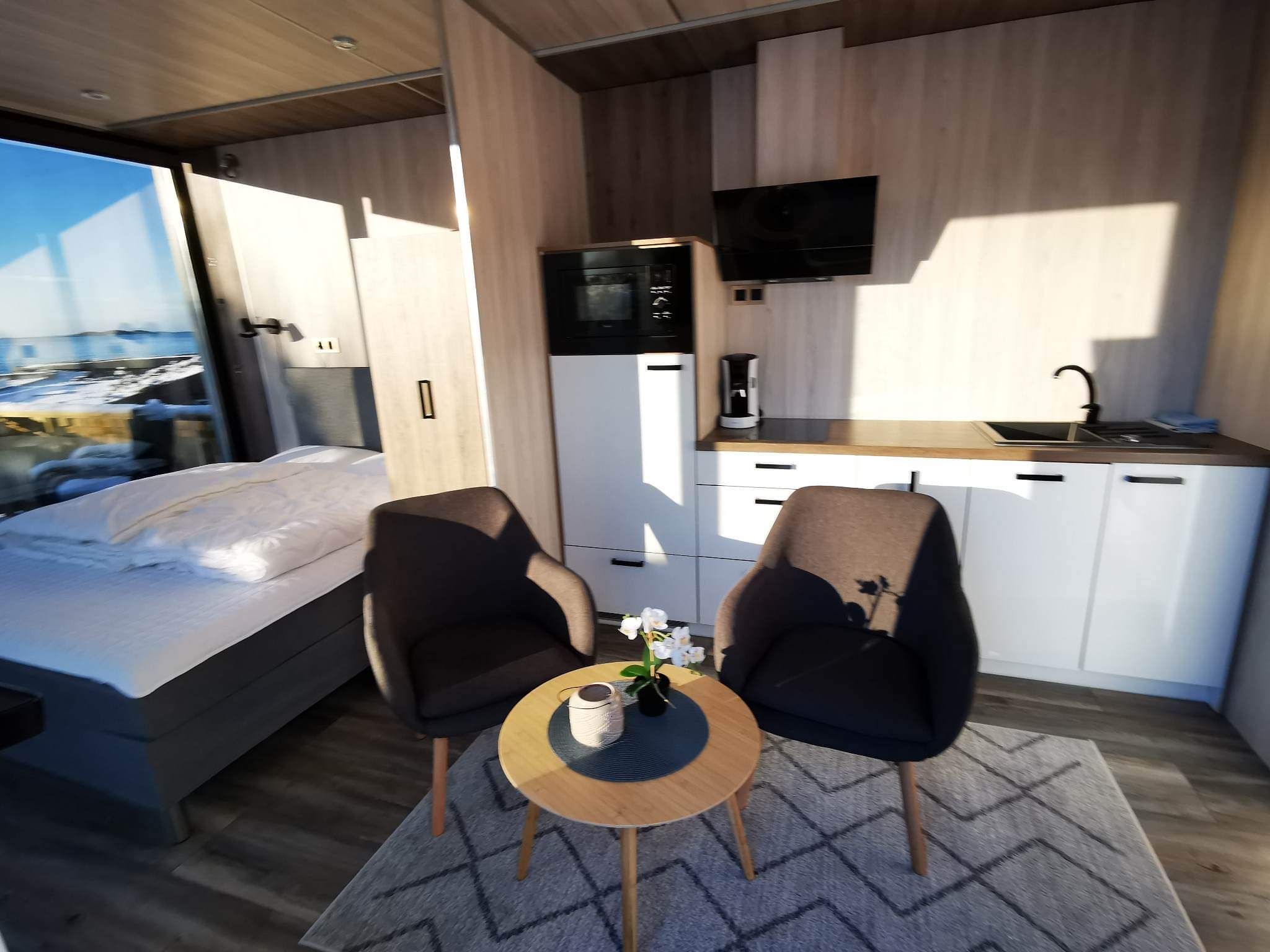 Habitación pequeña con cama, dos sillas, una mesita con un florero, microondas, cafetera y televisor montado en la pared.