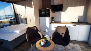 Habitación pequeña con cama, dos sillas, una mesita con un florero, microondas, cafetera y televisor montado en la pared.
