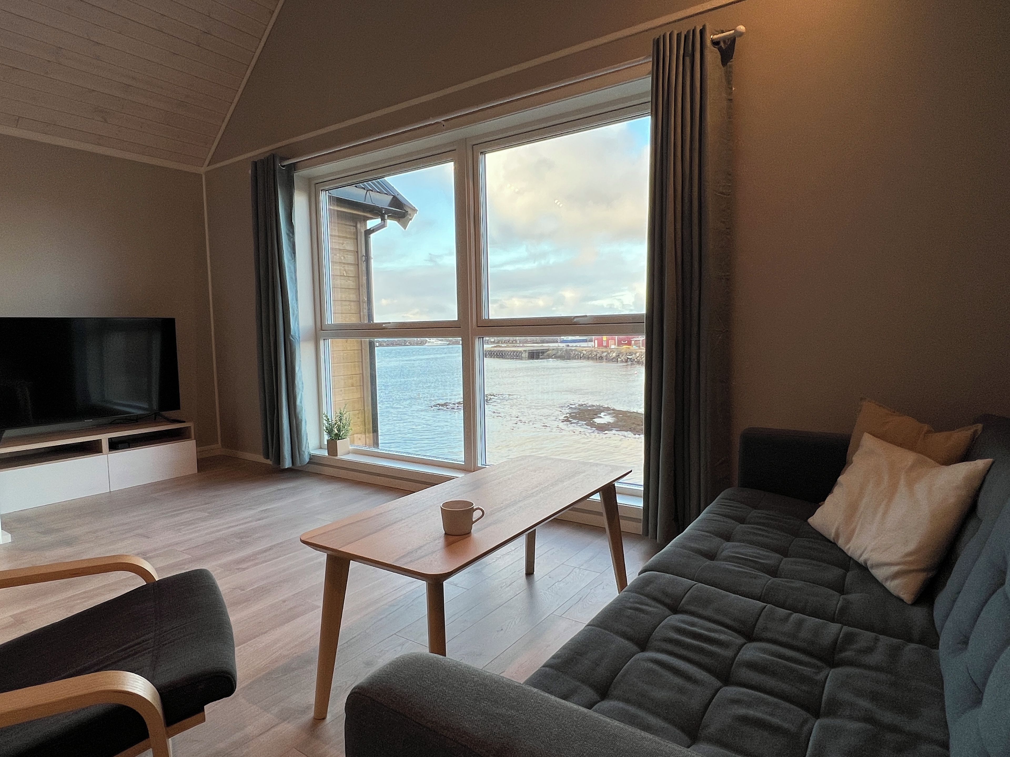 A living room with a television, a sofa, a table, and a view of a body of water through large windows.