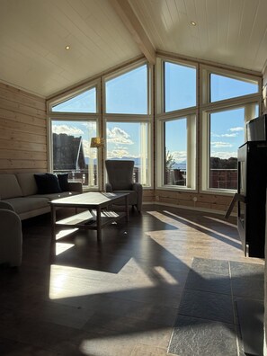 A room with a sofa, a table, and a chair, large windows with a view of the sky and mountains, and a ceiling with wooden beams.