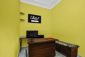Bancada de recepção com uma placa em cima, um monitor de computador, uma mesa pequena, uma cadeira e um banco.