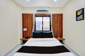 A hotel room with a bed, a bedside table with a phone, a window with curtains, an air conditioning unit, and a framed picture on the wall. - Super Collection O MIT Alandi (Rajgurunagar)
