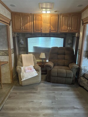 A cozy RV interior with a large window, wooden cabinets, two reclining chairs, and a small table with a lamp. - Charming RV Stay on a Working Farm-Ultimate Country Escape! (Cottondale)