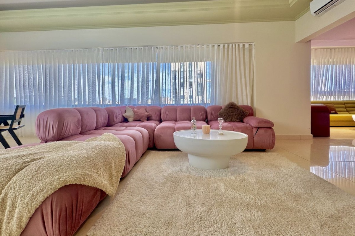 A modern living room with a large sectional sofa, a round coffee table, and a large window with sheer curtains.