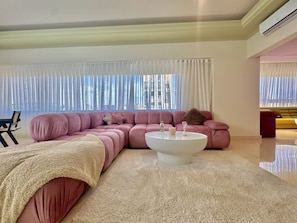 A modern living room with a large sectional sofa, a round coffee table, and a large window with sheer curtains. - Riverside Residences by Stayinn (Sin El Fil)