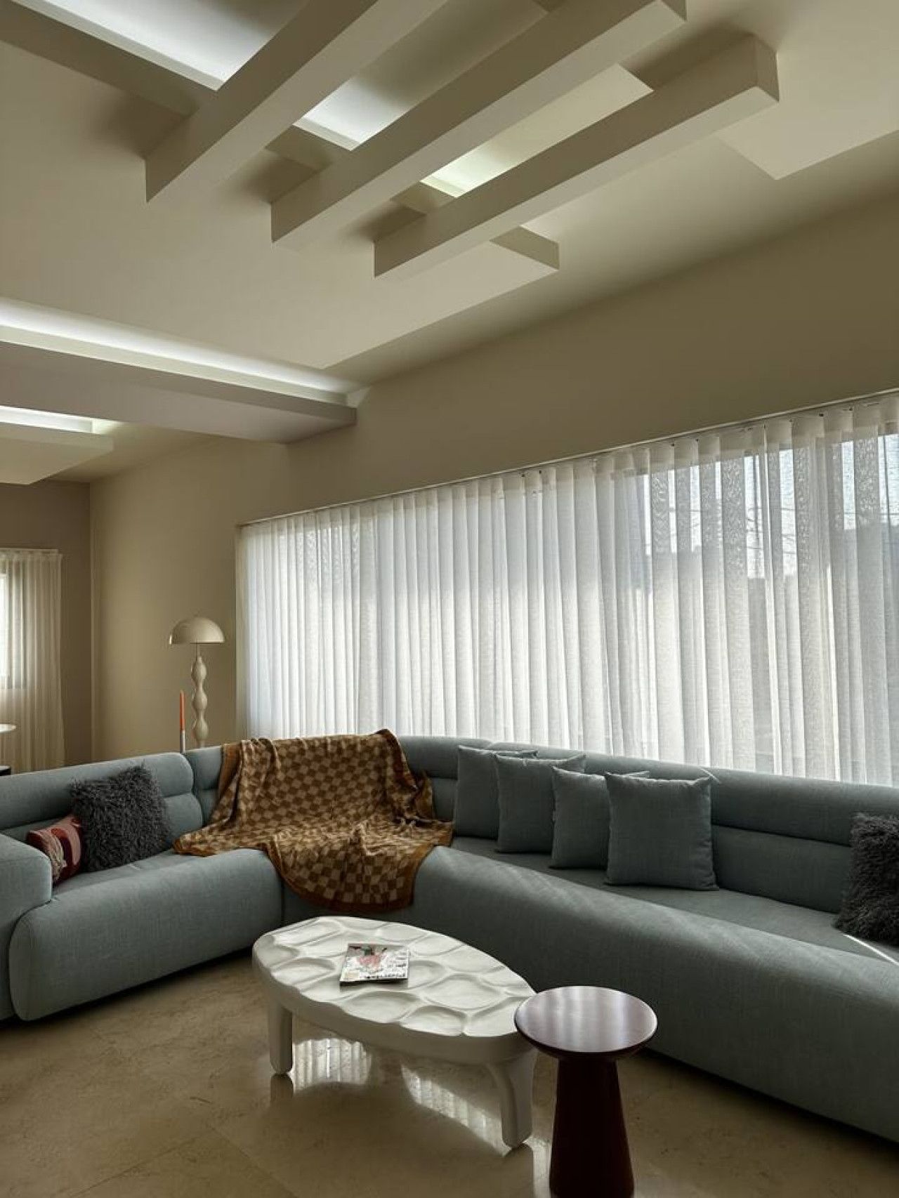 A modern living room with a sectional sofa, a coffee table, and a large window with sheer curtains.