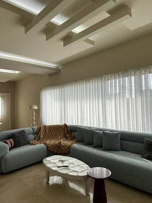 A modern living room with a sectional sofa, a coffee table, and a large window with sheer curtains. - Riverside Residences by Stayinn (Sin El Fil)