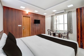 Una habitació d'hotel amb un llit gran, un televisor fixat a la paret, una taula petita amb cadires i una finestra amb cortines.