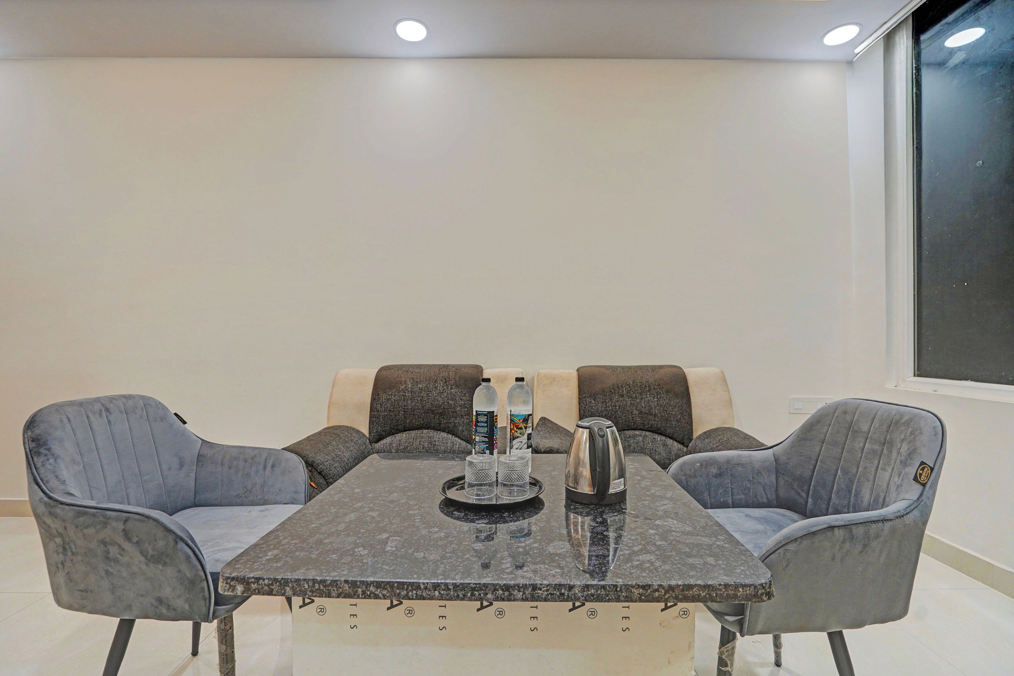 A conference room with a rectangular table, four grey upholstered chairs, a water bottle, a glass, and a kettle.