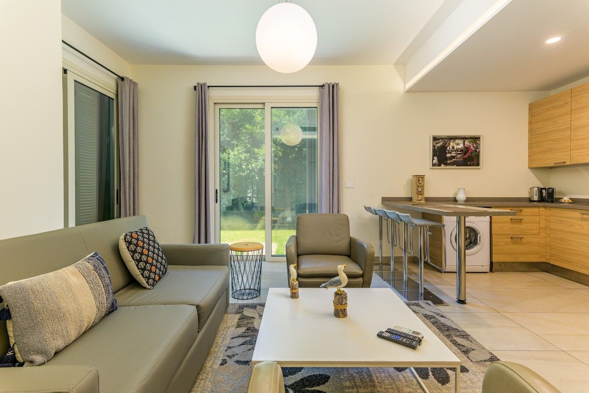 Una sala moderna con sofá, sillón y mesa de centro, un área de cocina con taburetes de bar y un ventanal corredero al exterior.