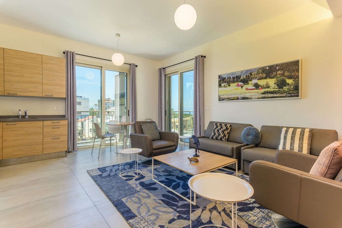 Una sala de estar moderna con sofá, mesa de centro y sillones, una cocina con armarios de madera y ventanas grandes con cortinas.
