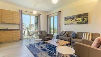 Una sala de estar moderna con sofá, mesa de centro y sillones, una cocina con armarios de madera y ventanas grandes con cortinas.