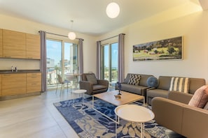 Una sala de estar moderna con sofá, mesa de centro y sillones, una cocina con armarios de madera y ventanas grandes con cortinas.