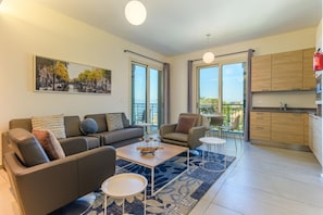 A modern living room with a sofa, armchair, coffee table, and a rug. There is a kitchen area with wooden cabinets and a red fire extinguisher. A large window provides a view of the outdoors.