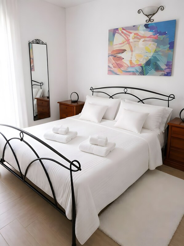 A neatly made bed with white linens and two rolled towels, a bedside table with a lamp, a wall-mounted mirror, and a colorful abstract painting above the bed.