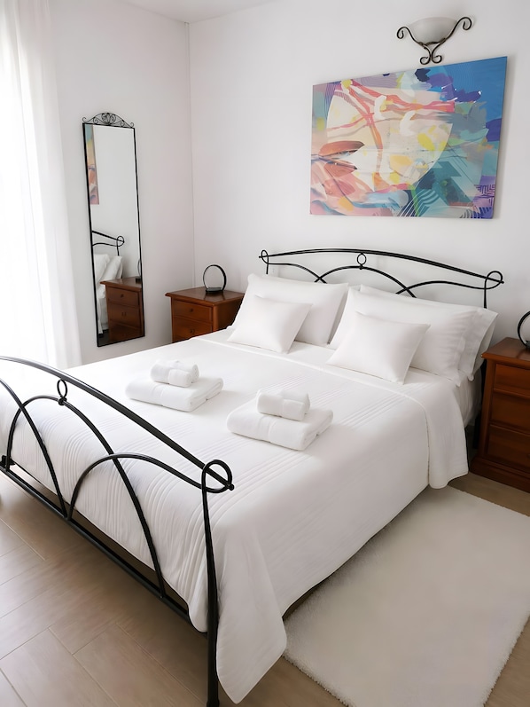 A neatly made bed with white linens and two rolled towels, a bedside table with a lamp, a wall-mounted mirror, and a colorful abstract painting above the bed.
