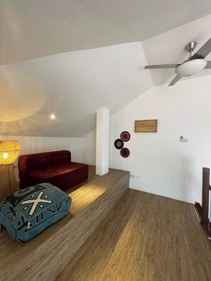 Una habitación con un sofá rojo, un reposapiés estampado, un ventilador de techo y un aplique de pared.