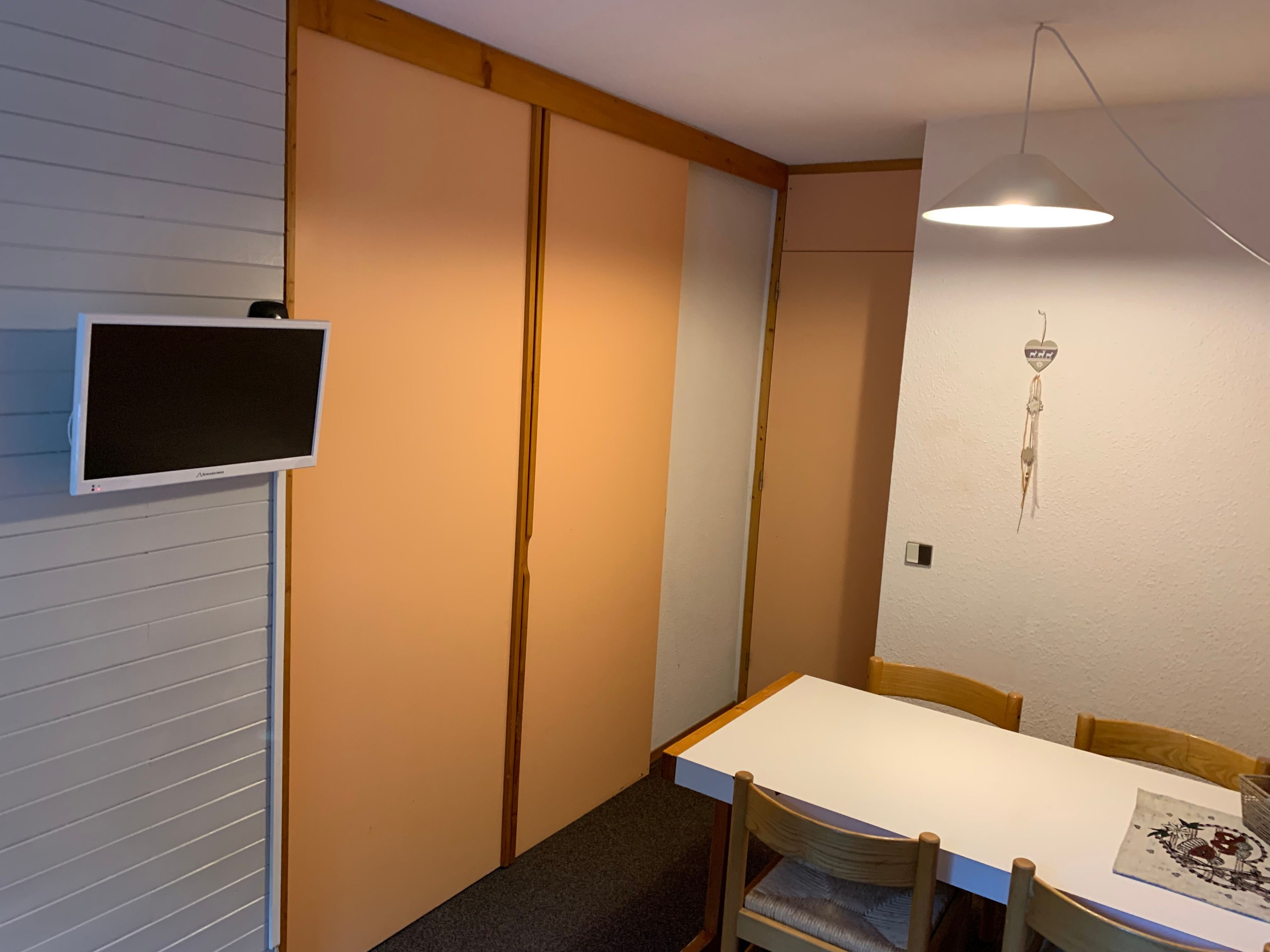 A room with a white table, wooden chairs, a TV mounted on the wall, and a hanging decoration.
