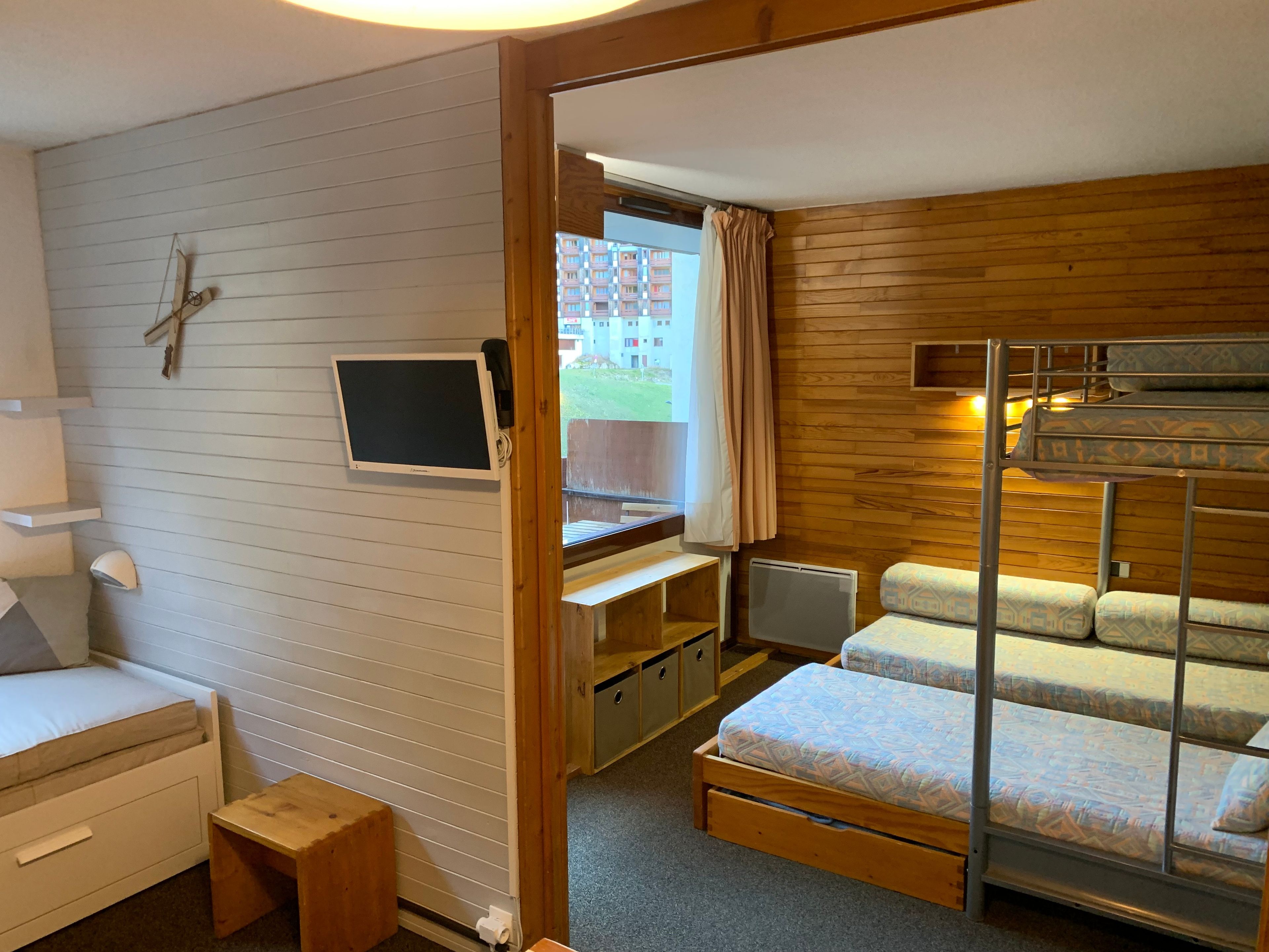Ein kleines Zimmer mit einem Etagenbett, einem Schreibtisch inklusive Computer, einem kleinen Nachttisch und einem Fenster mit Vorhängen.