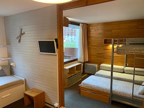 A compact room with a bunk bed, a desk with a computer, a small bedside table, and a window with curtains.
