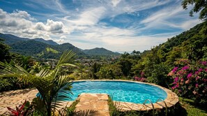 A swimming pool with a stone border, surrounded by lush greenery and mountains in the background. - New Listing (Paraty)