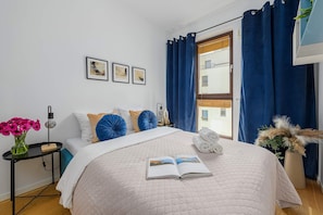 Een opgemaakt bed met een gewatteerde spreideken, een nachtkastje met een lamp, een vaas met bloemen en een raam met gordijnen.