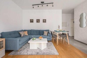 A modern living room with a blue sofa, a white coffee table, and a dining area with a white table and chairs. - Wola Flat with Free Parking (Warszawa)