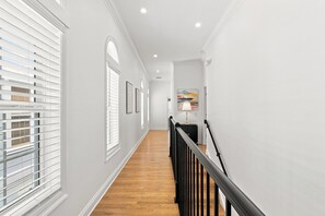 A hallway with a wooden floor, white walls, and a black railing. There are windows with blinds on the left and a painting on the right. - Welcome to Soleil House, a  Seacrest Beach retreat perfectly located on 30A. (Seacrest)