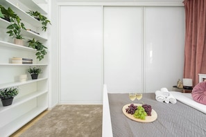 A modern room with a white wardrobe, a small table with a plate of grapes and two glasses of white wine, a beige carpet, and a pink cushion on a bench. - Apartment near River Walks (Warszawa 2)